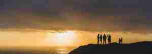 small family standing on cliff looking out towards the sun setting over the water