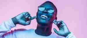 Young african man listening to music with earphones, dancing isolated on pink background