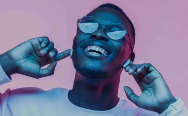 Young african man listening to music with earphones, dancing isolated on pink background