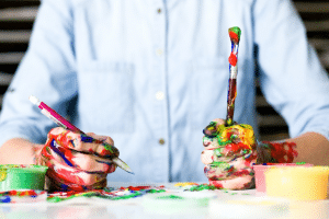 man in blue shirt holding pen and paintbrush with hands covered in red, green and yellow paint